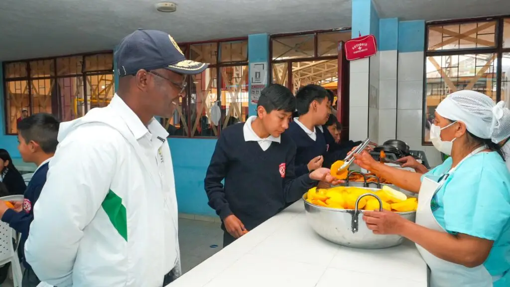 Una manipuladora de alimentos con cofia, tapabocas y delantal sirve fruta a un estudiante en la barra de atención mientras un supervisor observa el proceso en el comedor escolar.