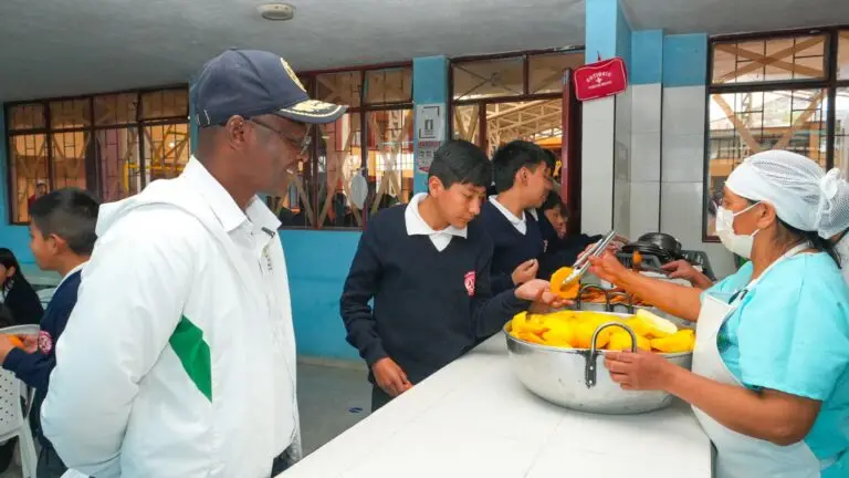 Una manipuladora de alimentos con cofia, tapabocas y delantal sirve fruta a un estudiante en la barra de atención mientras un supervisor observa el proceso en el comedor escolar.