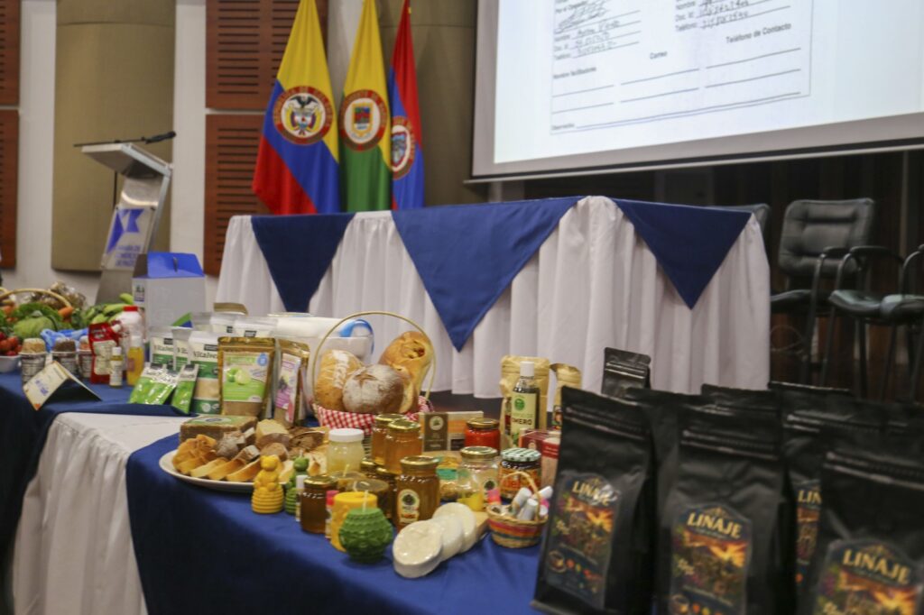 Una mesa de visualización con productos alimenticios, incluyendo tomates frescos, cebollas y verduras en el extremo izquierdo. Panes en una canasta y paquetes de panela en bolsas de papel se sientan en el medio. Frascos de miel y velas en el primer plano central. Paquetes negros de café "LINAJE" en primer plano a la derecha. Un banner de fondo tiene texto de formulario escrito a mano.
