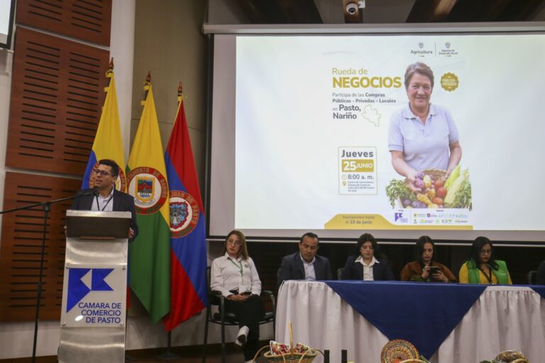 Un hombre con gafas de pie detrás de un atril que tiene el logo de la CAMARA DE COMERCIO DE PASTO. Hay tres banderas a la izquierda (la bandera colombiana y dos banderas de Nariño) y un panel de personas sentadas en una mesa en primer plano. Un proyector en la pared muestra el póster del evento "RUEDA de NEGOCIOS".