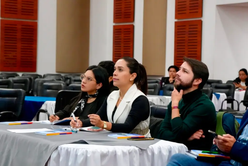 Tres asistentes, dos mujeres y un hombre, prestan atención y toman notas con bolígrafos durante un taller técnico de la Gobernación de Nariño en un auditorio.