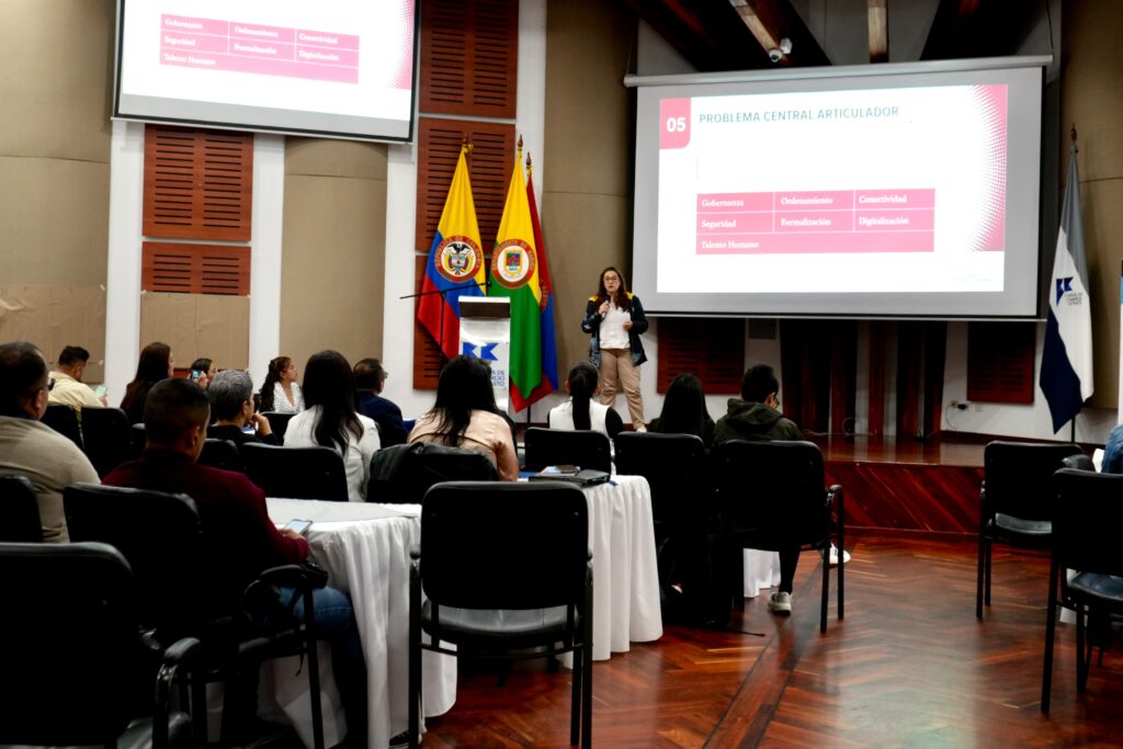 Una expositora dicta una conferencia frente a un auditorio en el salón de la Cámara de Comercio de Pasto. La gran pantalla proyecta una diapositiva titulada "Problema Central Articulador" con ejes como Gobernanza, Seguridad, Ordenamiento y Conectividad.