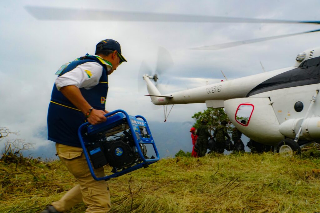 Personal de socorro de la Gobernación de Nariño transportando un generador eléctrico azul cerca de un helicóptero blanco en una zona montañosa y nublada.