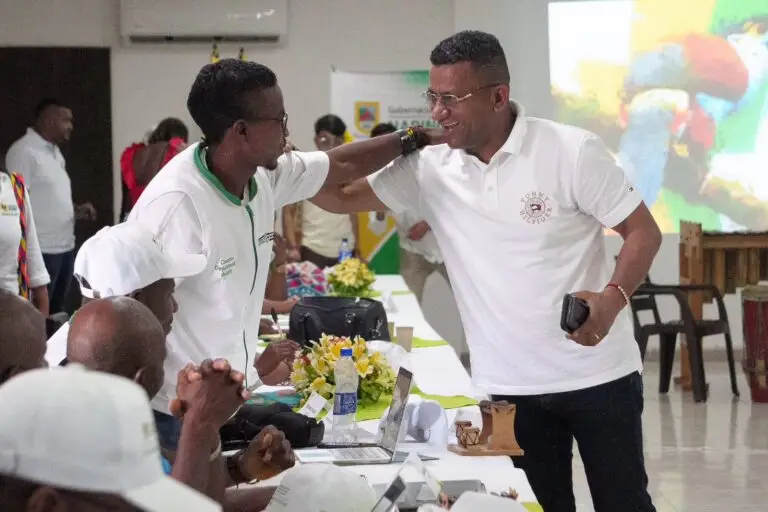 Un hombre afrodescendiente con gafas y polo blanca sonríe mientras saluda afectuosamente con la mano en el hombro a otro participante en un salón de eventos con mesas de trabajo.