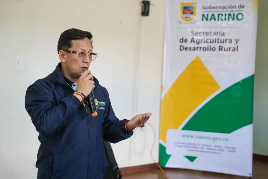 Funcionario con chaqueta azul de la Gobernación de Nariño y escarapelas institucionales dirigiéndose al público con un micrófono junto a un pendón de la Secretaría de Agricultura.