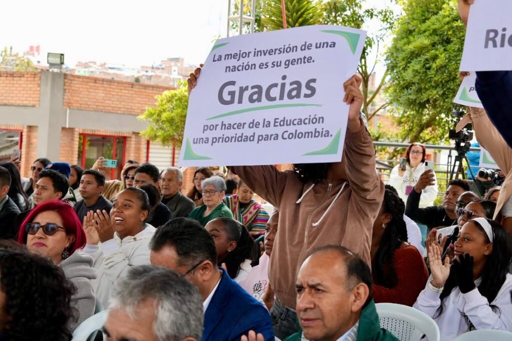 Asistentes al evento aplauden mientras una persona sostiene un cartel impreso en alto que dice "La mejor inversión de una nación es su gente. Gracias por hacer de la Educación una prioridad para Colombia".