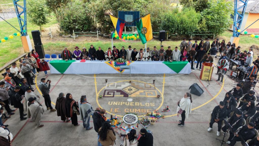Vista elevada de la mesa principal y el polideportivo de la Institución Educativa Cumbe, con una mandala ceremonial en el centro del suelo que dice "UNIPUEBLOS I.E. CUMBE", autoridades indígenas en la mesa y asistentes alrededor.
