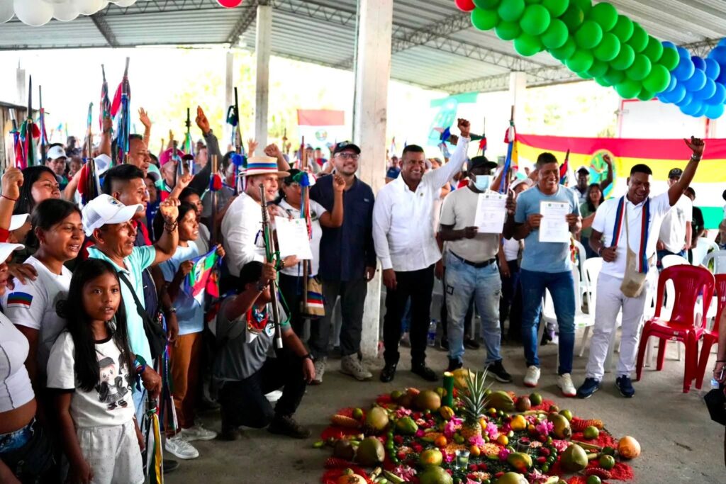 El gobernador Luis Alfonso Escobar Jaramillo, el director de la ANT Felipe Harman y líderes afrodescendientes e indígenas levantan los brazos celebrando en torno a una mandala de frutas y flores organizada en el suelo durante la entrega de predios en Tumaco.