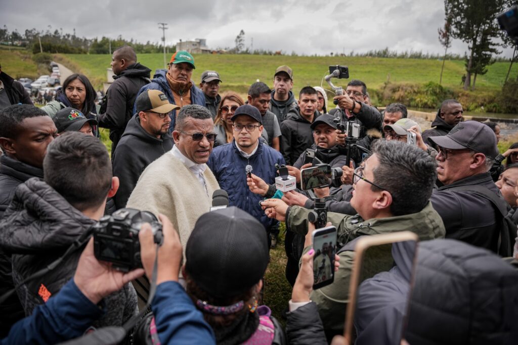 Gobernador de Nariño con ruana artesanal dando declaraciones rodeado de periodistas con micrófonos y cámaras en espacio abierto.