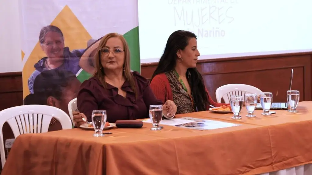 Liliana Montúfar, Secretaria de Equidad de Género e Inclusión Social, sentada en una mesa principal vestida con una blusa tono vinotinto, junto a una funcionaria, durante un evento institucional.