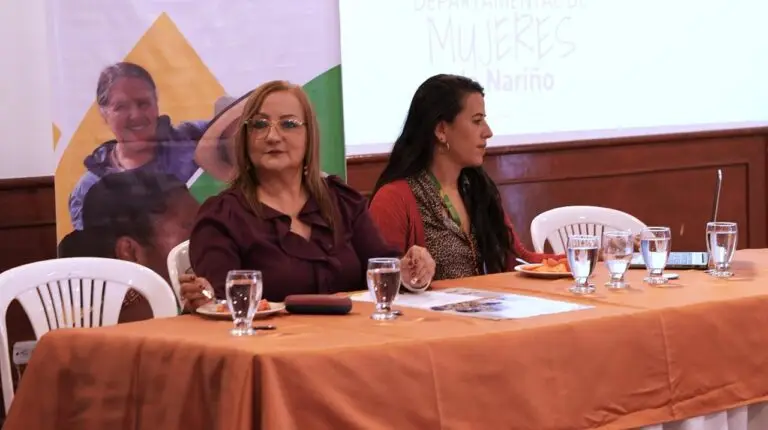 Liliana Montúfar, Secretaria de Equidad de Género e Inclusión Social, sentada en una mesa principal vestida con una blusa tono vinotinto, junto a una funcionaria, durante un evento institucional.
