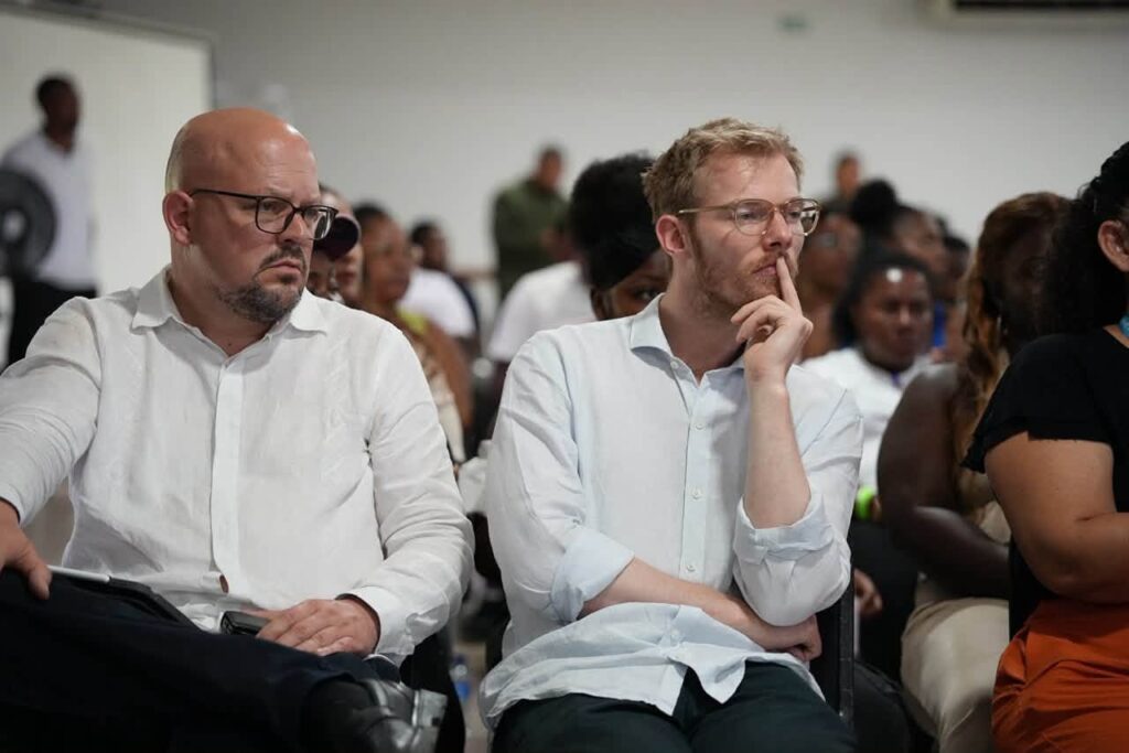 Dos delegados internacionales con camisa clara y gafas sentados atentos en el público durante un evento institucional en auditorio.