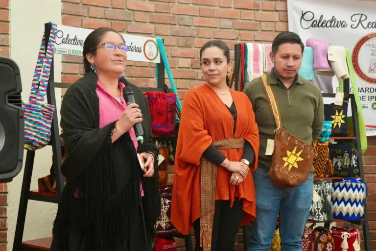 Una artesana indígena con chal negro y gafas habla por un micrófono junto a Claudia Patricia Erazo Casabón, Directora de Turismo, quien viste una ruana anaranjada con faja artesanal, frente a un exhibidor de bolsos tejidos.