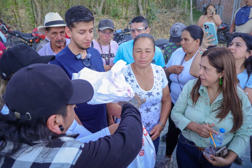 Comunidad y funcionarios reunidos alrededor de la entrega de sacos de ayuda en Ancuya, observados por una mujer con camisa verde menta que toma apuntes.