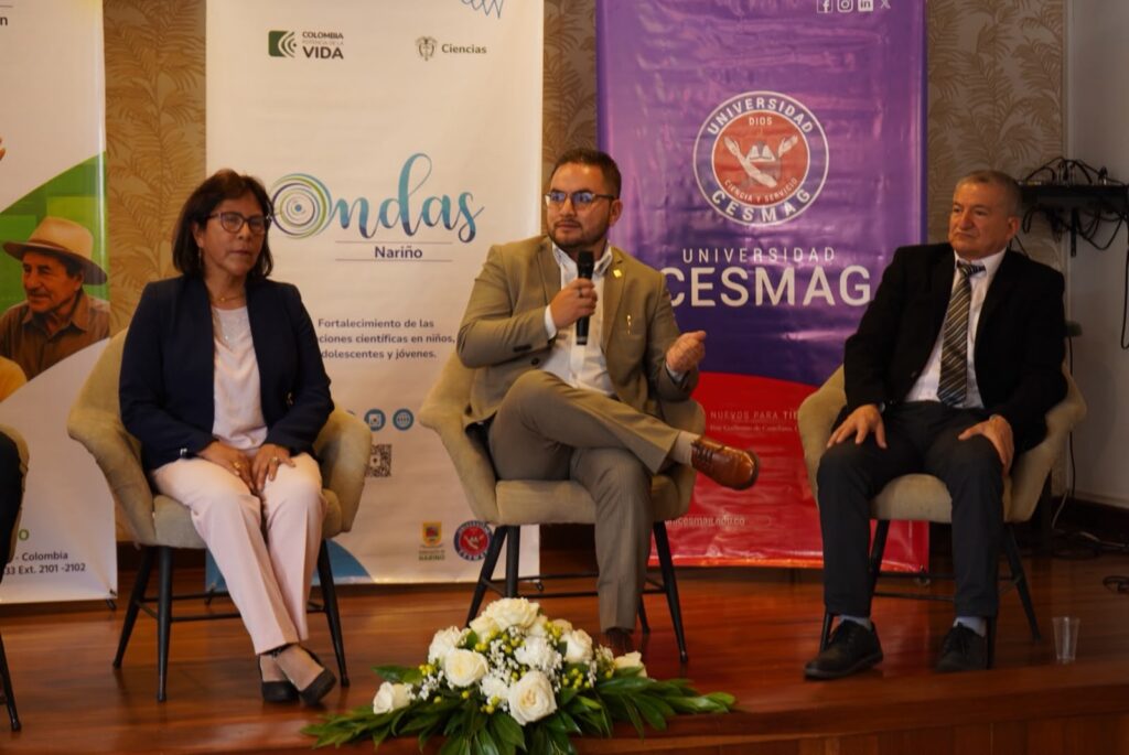 Panel de tres personas sentadas en una tarima durante un evento formal, con un hombre en el centro hablando por un micrófono, flanqueado por pendones institucionales de Ondas Nariño, Minciencias y la Universidad CESMAG.
