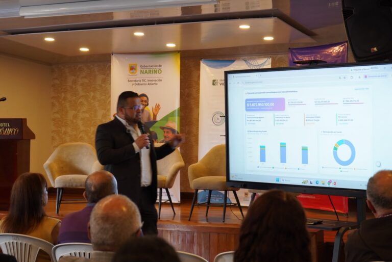 Funcionario hablando por micrófono frente a un auditorio y apuntando a una pantalla digital interactiva que muestra el valor "$5.672.950.100,00" junto a gráficos de barras y circulares, al lado de un pendón de la Secretaría TIC, Innovación y Gobierno Abierto.