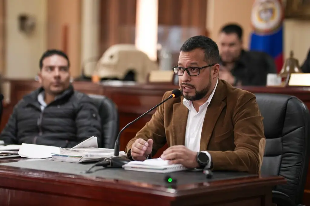 Hombre con gafas y saco café hablando por un micrófono en un escritorio del recinto de la Asamblea Departamental.
