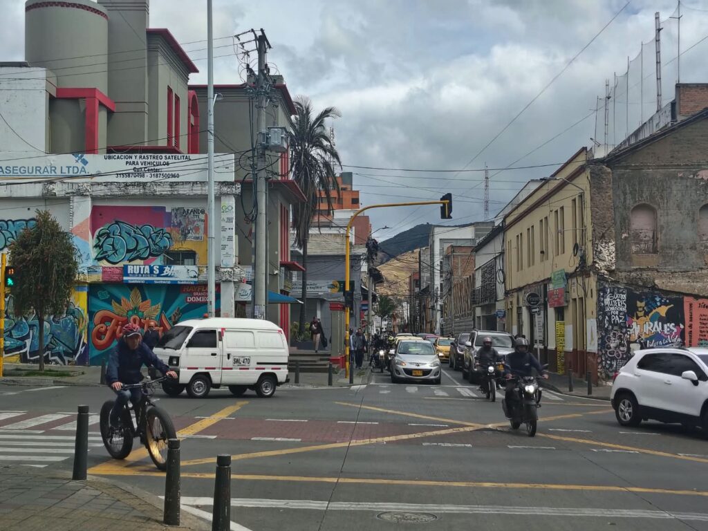 Tránsito variado de ciclistas, motociclistas, automóviles y vans en una intersección vial urbana en Nariño.