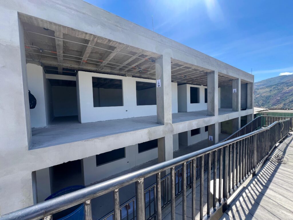 Estructura de concreto de dos pisos en construcción, con espacios abiertos para ventanas y pasillos delimitados por barandas metálicas negras, correspondiente a la nueva sede de la Institución Educativa Rafael Uribe Uribe.