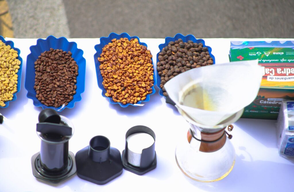 Diferentes niveles de tostión de granos de café presentados en recipientes azules junto a métodos de preparación alternativos sobre una mesa blanca.