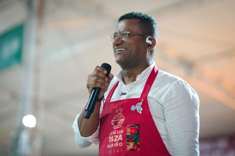 El Gobernador de Nariño sonriendo y hablando por un micrófono, vistiendo una camisa blanca y un delantal rojo con el logo de "La Mejor Taza Nariño 2026".