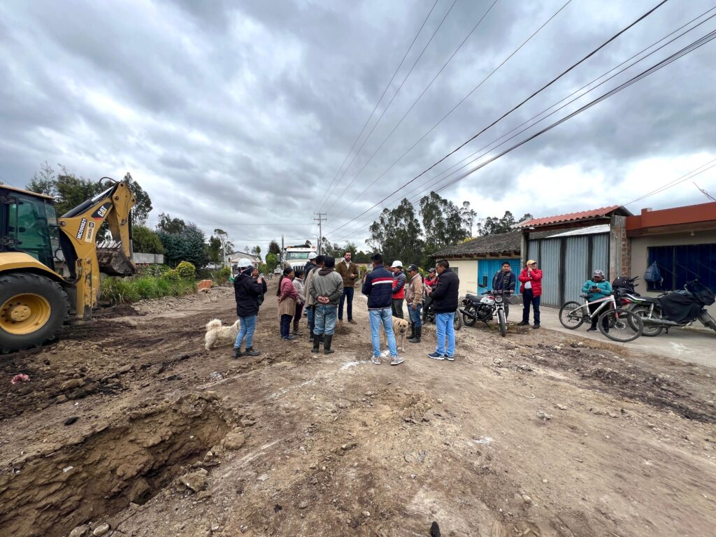 Grupo de habitantes y personal técnico con cascos blancos reunidos sobre una vía en construcción, junto a una retroexcavadora amarilla y una zanja abierta en la tierra.