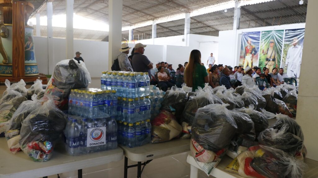 Mesas apiladas con pacas de botellas de agua y grandes bolsas transparentes que contienen mercados, ropa y elementos de primera necesidad, con un grupo de personas reunidas al fondo en un amplio salón.