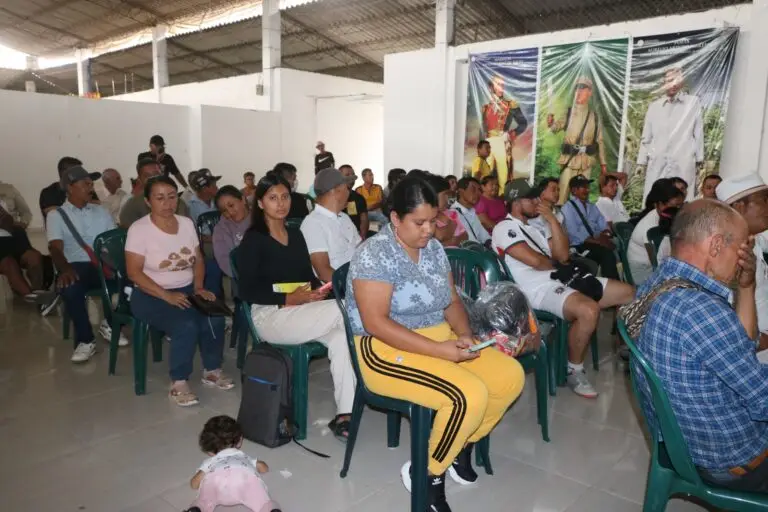 Un grupo de hombres y mujeres de distintas edades sentados en sillas plásticas verdes dentro de un recinto amplio, escuchando atentamente durante la jornada de entrega de ayudas; al fondo se observan pendones con figuras históricas.