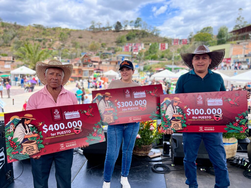 Tres productores cafeteros sonrientes sostienen cheques gigantes de premiación por el primer, segundo y cuarto puesto en la categoría Artesanal Tradicional de La Mejor Taza Nariño 2026, durante un evento al aire libre en El Peñol.