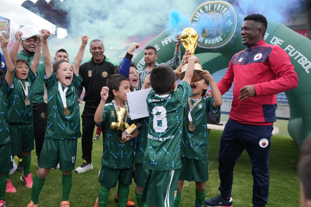 Niños del equipo Deportivo Pasto celebrando con euforia al levantar el trofeo del Mundialito Infantil de Fútbol de La Paz Territorial, acompañados por adultos y bajo humo azul de celebración en el estadio.