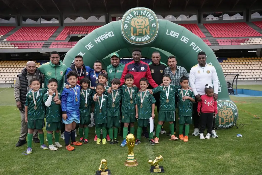 Fotografía grupal del equipo infantil campeón con uniformes verdes y medallas, posando junto a sus entrenadores y autoridades en la cancha del estadio, con trofeos dorados en primer plano.