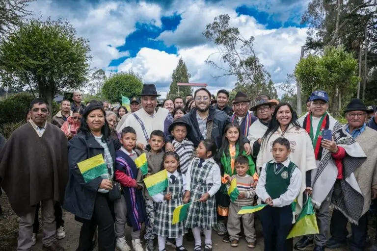 Representantes del gobierno, líderes indígenas y niños con uniformes escolares sosteniendo banderas verdes y amarillas, posando sonrientes al aire libre.