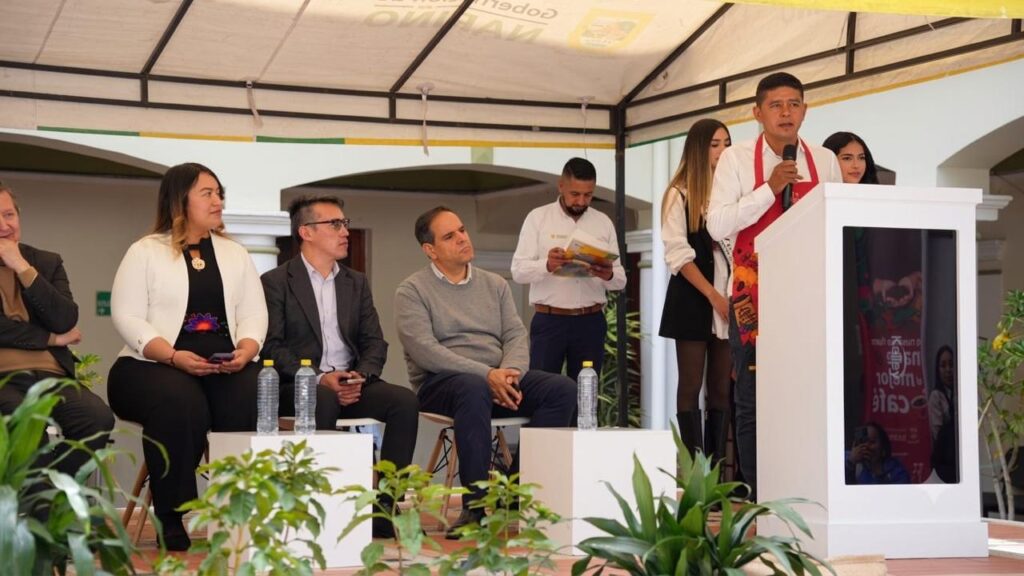 Hombre con delantal rojo hablando desde un atril blanco frente a un grupo de autoridades y panelistas sentados durante un evento oficial bajo una carpa.