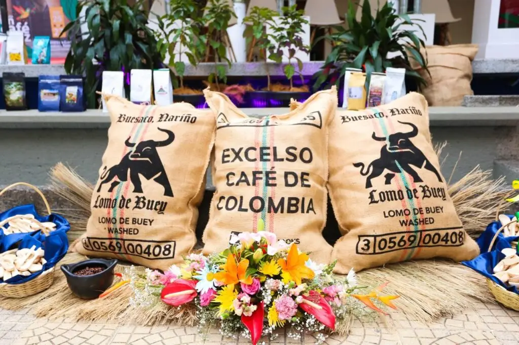 Exhibición de tres sacos de yute de café estampados con las marcas "Lomo de Buey, Buesaco, Nariño" y "Excelso Café de Colombia", decorados con un arreglo floral y pequeñas canastas.