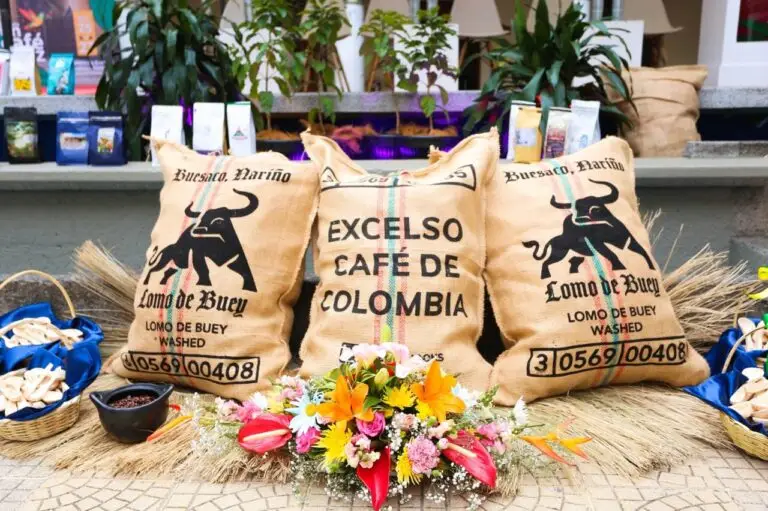 Exhibición de tres sacos de yute de café estampados con las marcas "Lomo de Buey, Buesaco, Nariño" y "Excelso Café de Colombia", decorados con un arreglo floral y pequeñas canastas.