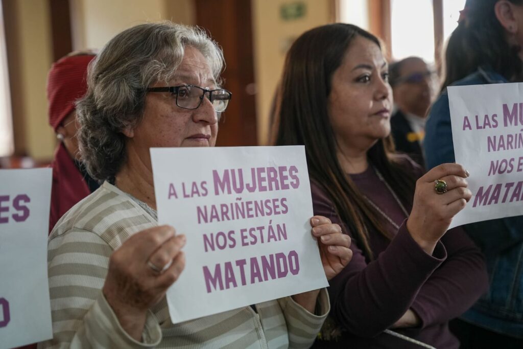 Una mujer mayor, de cabello canoso y gafas, con expresión de dolor y determinación, sostiene un cartel blanco que dice con letras moradas: "A LAS MUJERES NARIÑENSES NOS ESTÁN MATANDO". Sus manos están curtidas por el.
