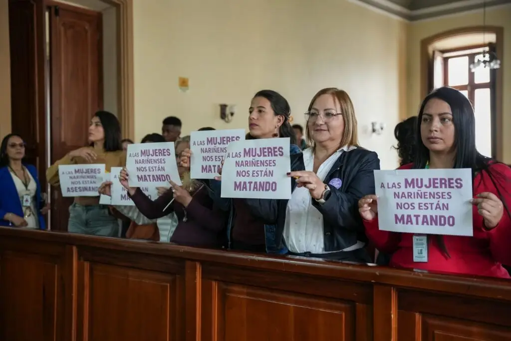 Un grupo diverso de mujeres se encuentra en el recinto de la Asamblea Departamental de Nariño, de pie detrás de una baranda de madera. Varias de ellas sostienen carteles blancos con la frase "A LAS MUJERES NARIÑENSES NOS ESTÁN MATANDO" en letras moradas. Entre ellas se encuentran la secretaria de Equidad de Género e Inclusión Social, Liliana Montúfar, y la diputada Isabel Rodríguez Guevara. El ambiente es de unidad y rechazo a la violencia.