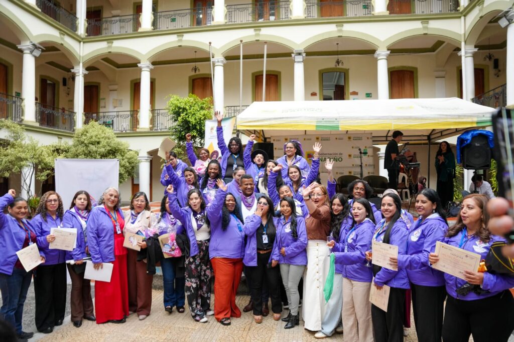 Grupo de mujeres diversas vistiendo chaquetas moradas y sosteniendo diplomas, celebrando junto al Gobernador de Nariño en el patio central de la Gobernación.