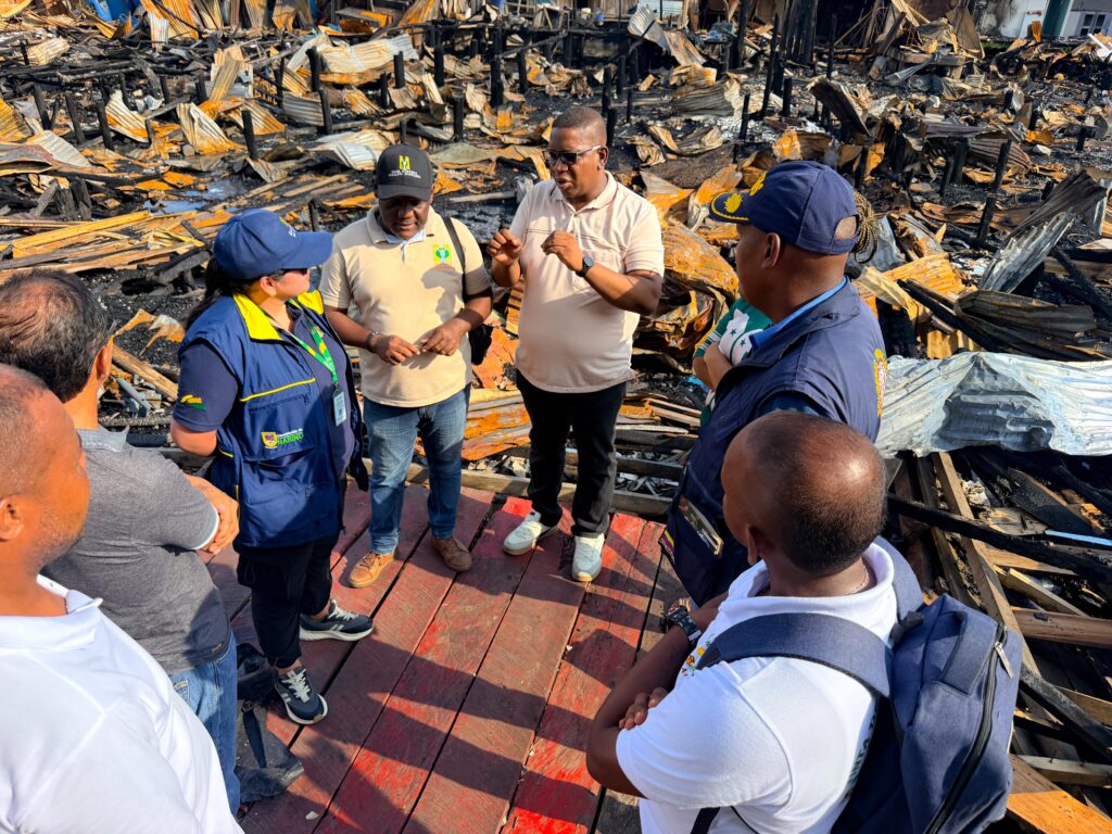 Grupo de autoridades departamentales y municipales reunidas sobre una pasarela de madera, rodeadas por una extensa zona de escombros, cenizas y láminas de zinc quemadas tras un incendio.v