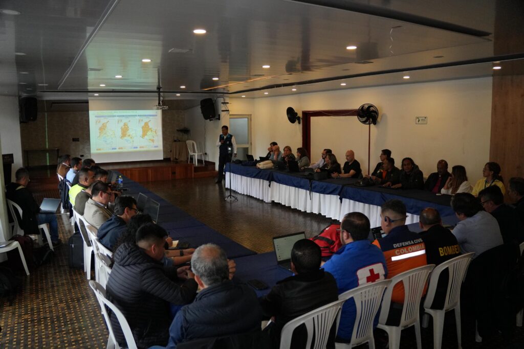 Reunión institucional en un salón amplio donde representantes del gobierno y miembros de organismos de socorro, como Defensa Civil y Bomberos, observan una presentación en video beam.