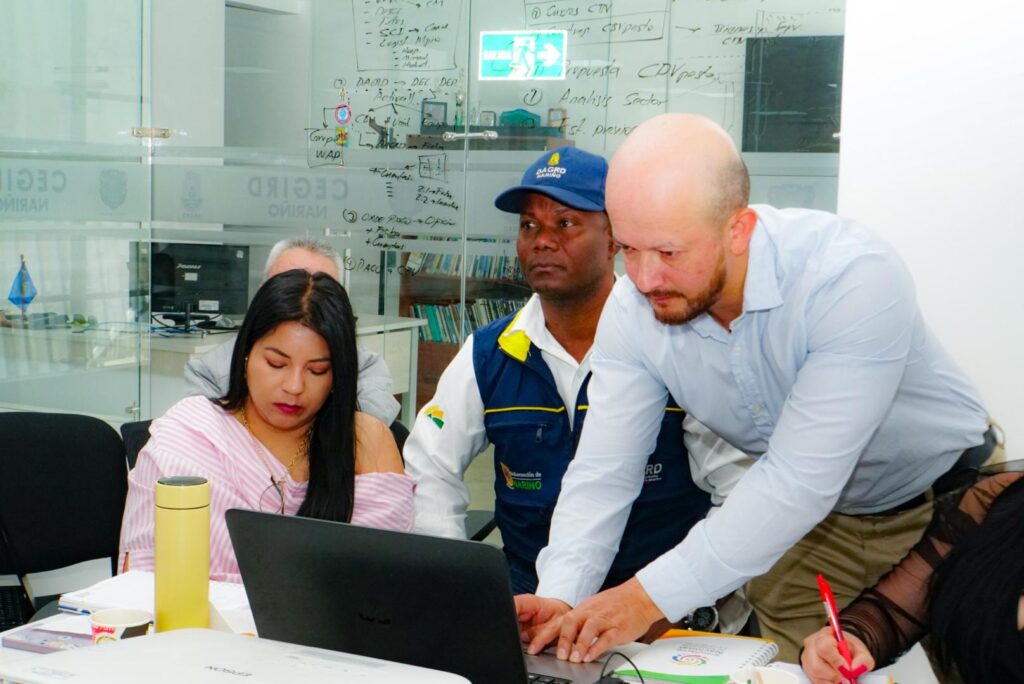 Tres funcionarios en una oficina revisando información en un computador portátil frente a un tablero de cristal con apuntes logísticos e institucionales.
