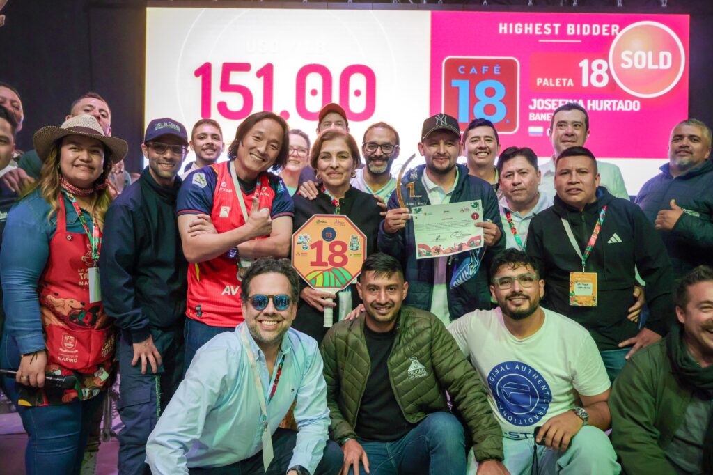 Grupo de personas celebrando frente a una pantalla gigante que muestra la cifra récord de 151.00 dólares y el logo de Café 18 durante un evento de subasta.