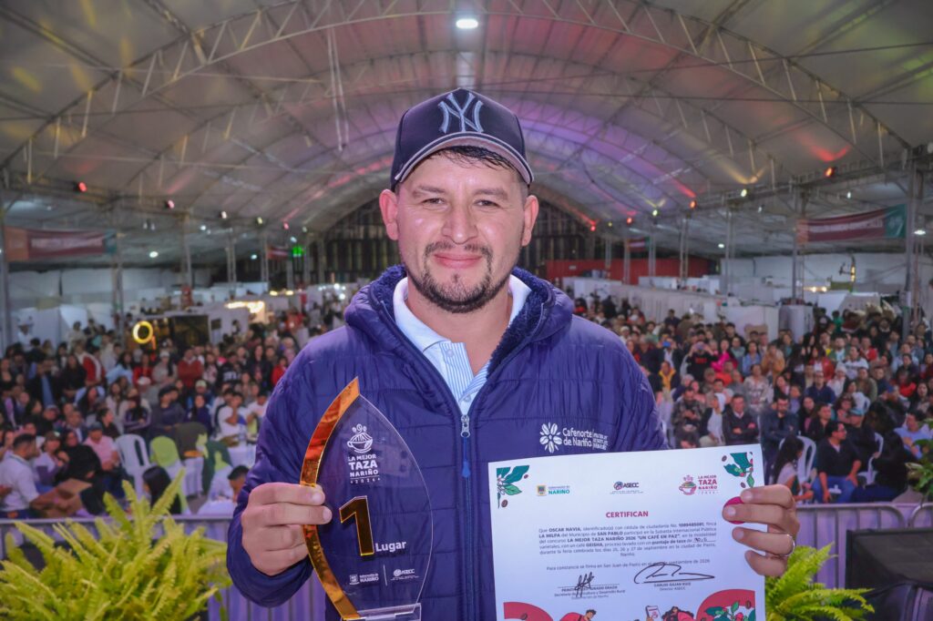 Hombre sonriente sosteniendo un trofeo de primer lugar y un certificado oficial del evento La Mejor Taza Nariño, con una gran multitud de asistentes sentados al fondo.
