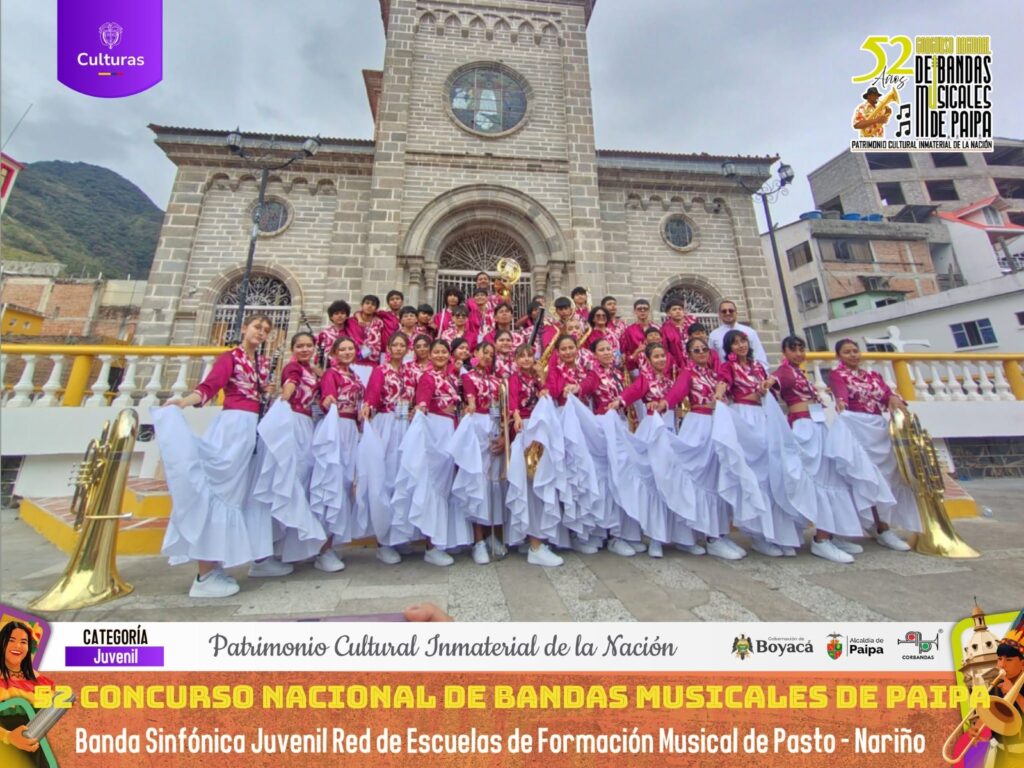 Integrantes de la Banda Sinfónica Juvenil de la Red de Escuelas de Formación Musical de Pasto vestidos con trajes folclóricos rosados y blancos posando frente a la iglesia de Paipa durante el 52° Concurso Nacional de Bandas.