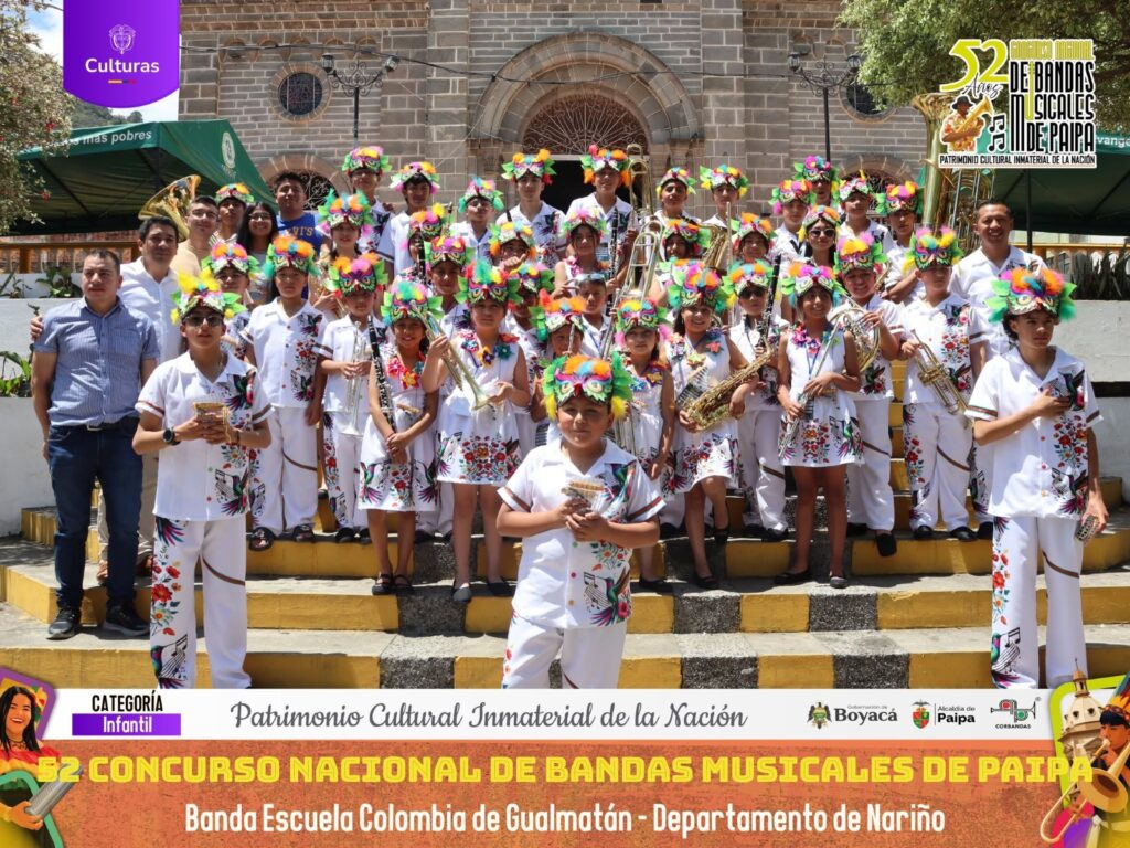 Niños integrantes de la Banda Escuela Colombia de Gualmatán con trajes típicos blancos bordados e insignias florales posando con sus instrumentos en Paipa