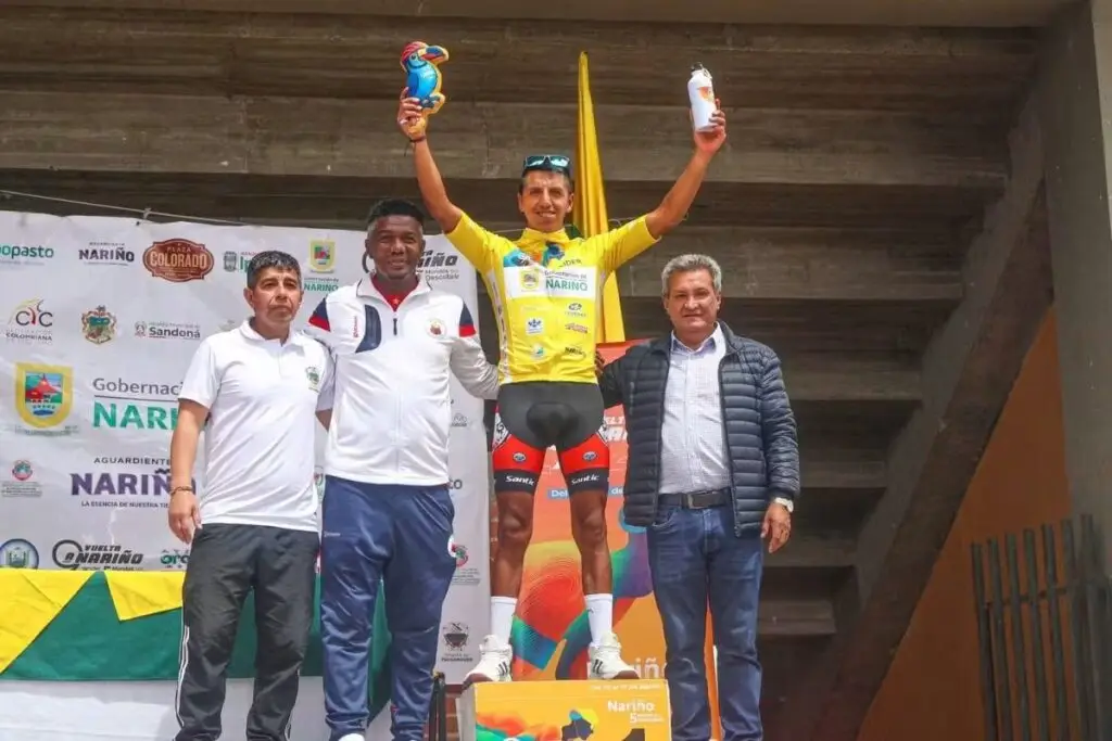 Ciclista vistiendo la camiseta amarilla de líder en lo alto de un podio, levantando un trofeo artesanal y una caramañola, acompañado por tres autoridades frente a un pendón institucional que incluye el logo de la Alcaldía de Sandoná.