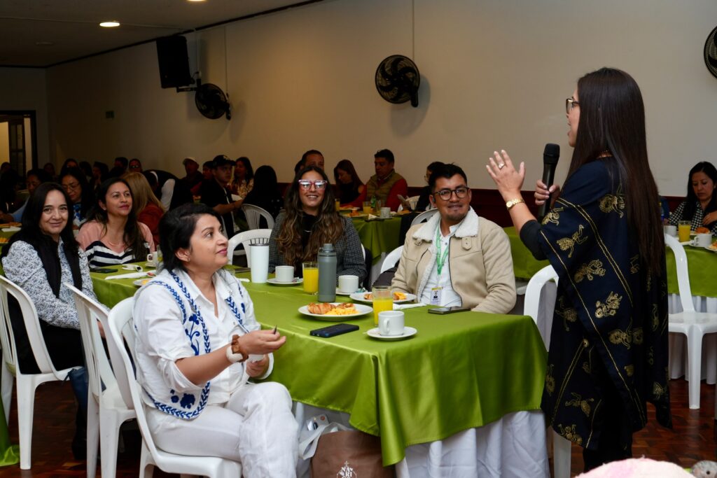 Mujer de pie hablando por un micrófono frente a un grupo de personas sentadas en mesas con manteles verdes.