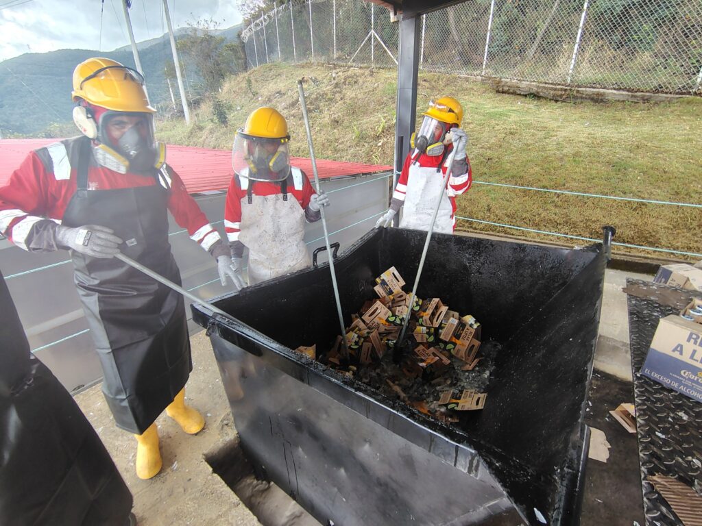 Operarios equipados con trajes de protección, cascos amarillos y máscaras triturando botellas de licor e inhabilitando productos de contrabando en un contenedor metálico en el relleno sanitario Antanas.