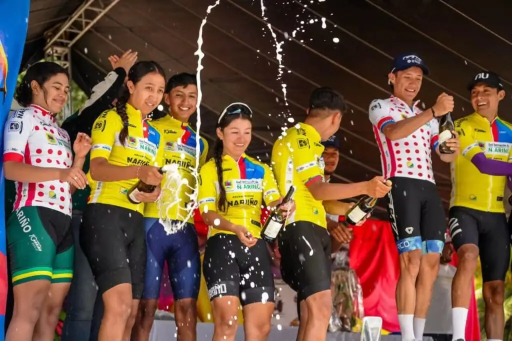Ciclistas de la Décima Vuelta a Nariño celebran en el podio durante la premiación, con uniformes de sus equipos y botellas de champaña.