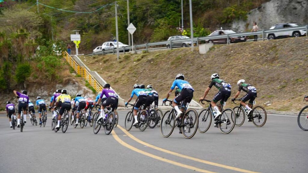 Pelotón de ciclistas transitando en curva sobre una vía asfaltada rodeada de vegetación y montañas durante la segunda etapa de la Vuelta a Nariño.